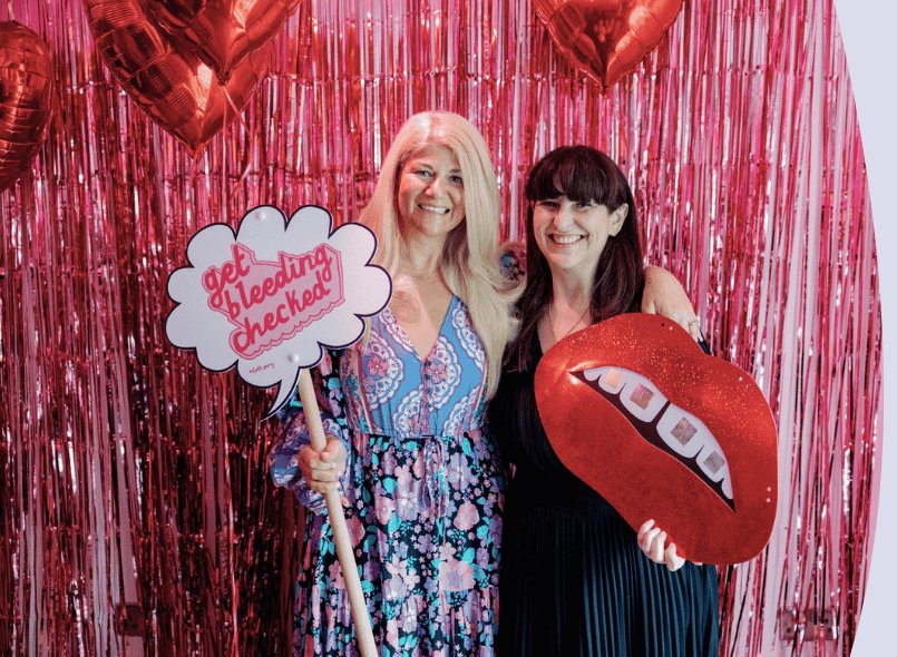 Two people with signs smiling with a pink backdrop. Sign says 'get bleeding checked'