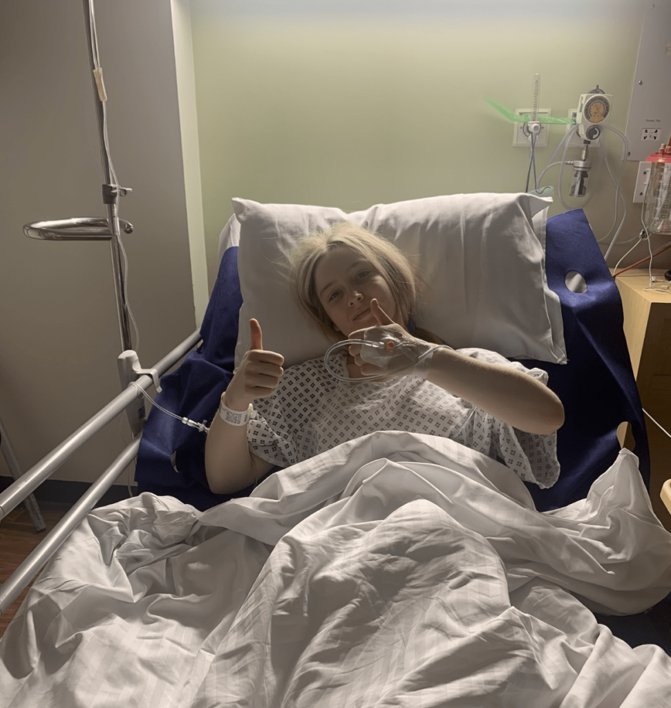 Picture of Pip lying in a hospital bed with her thumbs up