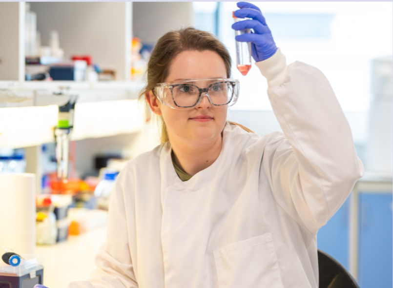 A photo of a scientist in a lab with their lab coat on holding up a test tube
