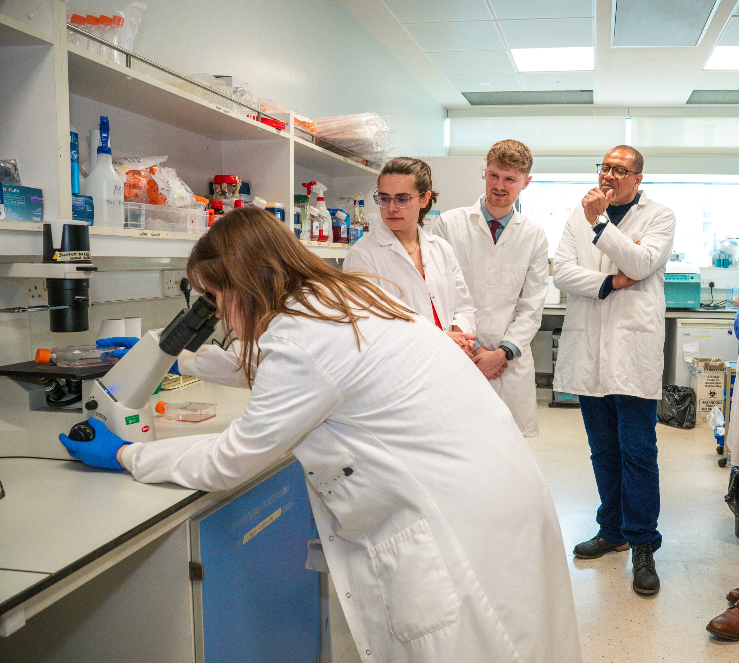 a group of people stood in the lab around someone looking down a microscope