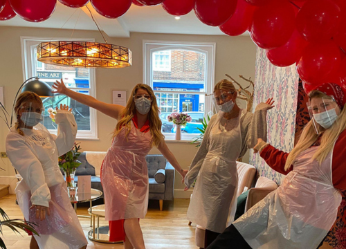 Photo of four people with arms outstretched, looking at camera. They are wearing face masks and lots of red balloons are around them.