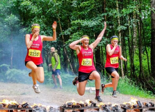 Photo of three people running and jumping over some flames on the ground. All wearing sweatbands, Eve Appeal tshirts and shorts. Two are holding their noses and has a hand over their arms in the air. 